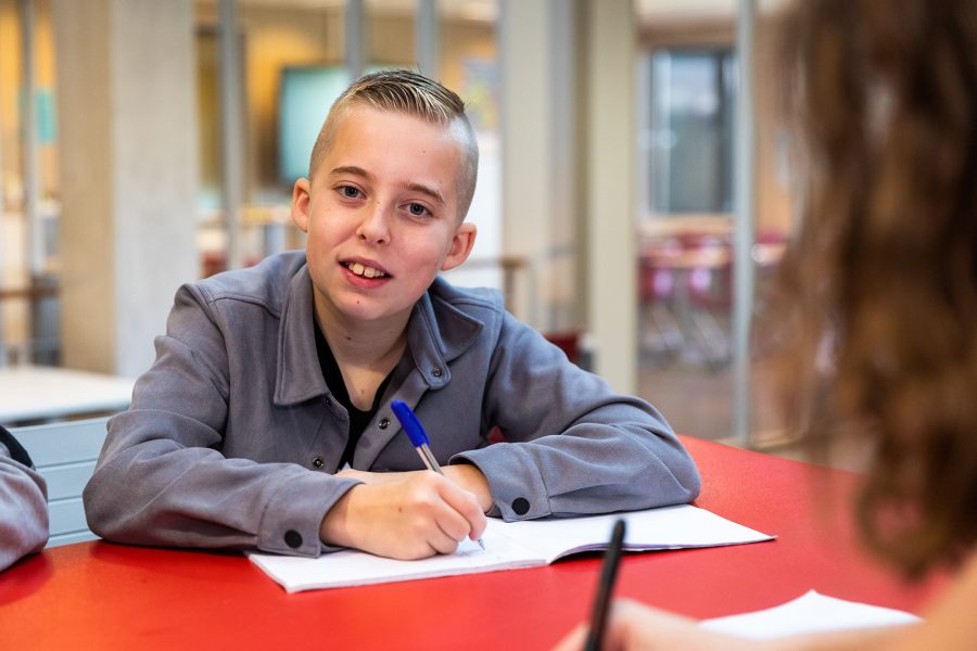 Jongen leerling van Houtens achter een bureau kijkt in de camera met pen en schrift bij de hand